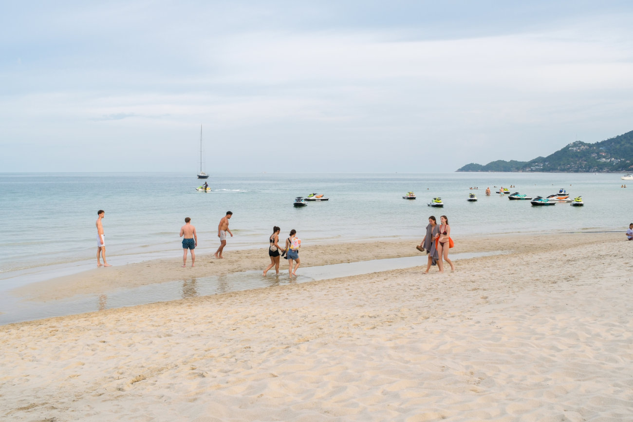 A group of people frolic on a beach in Koh Samui with jet skis and a yacht in the background. 